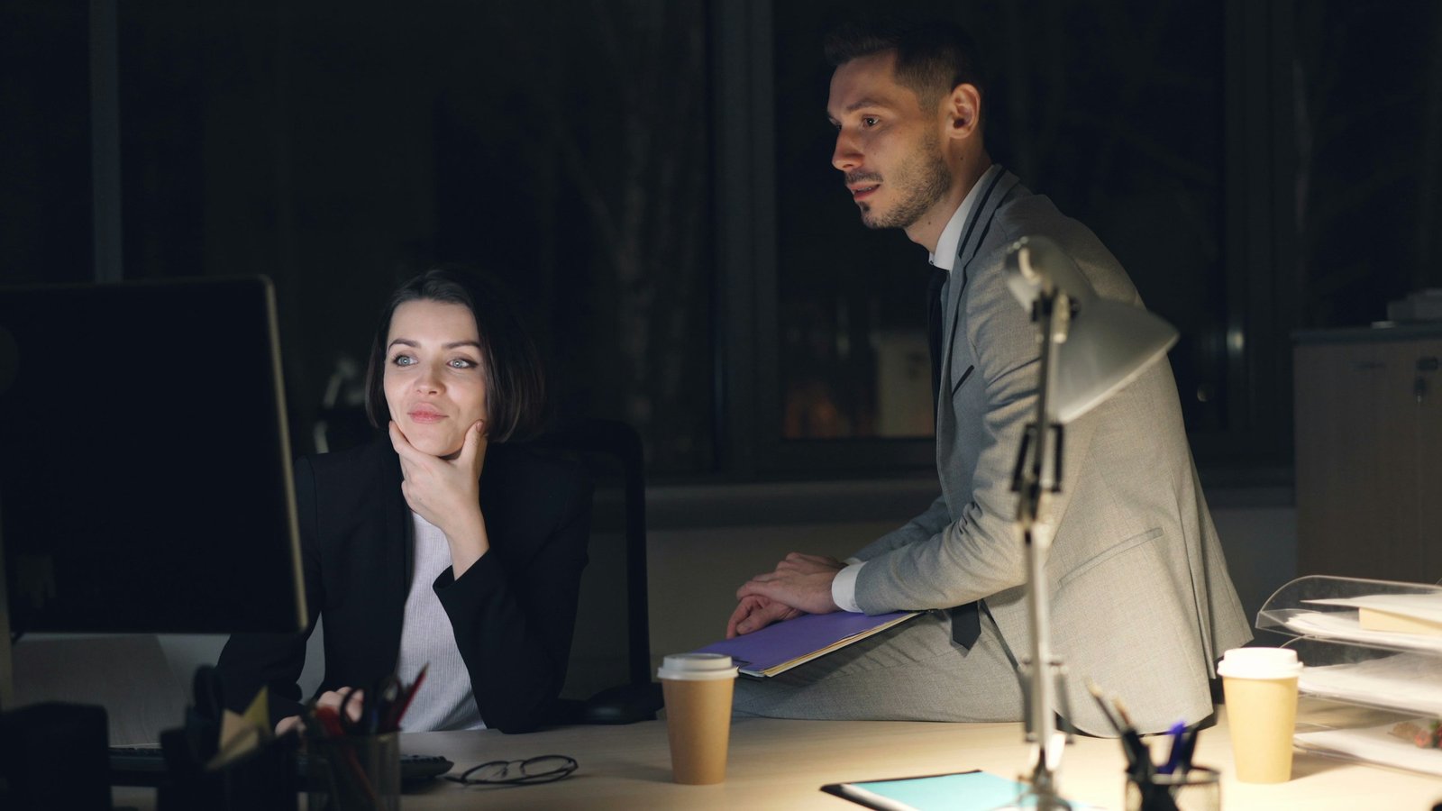 Marketing manager and a colleague working late in a dim office, waiting on a campaign landing page stuck in the dev queue